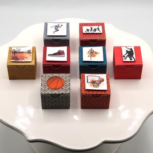 May include: A collection of nine small square gift boxes featuring various basketball-themed designs, including silhouettes of players, a basketball sneaker, a basketball, and a hoop, arranged in a grid on a white scalloped display stand.