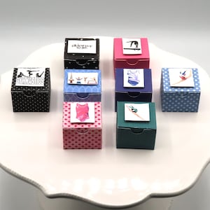 May include: A collection of eight small square gift boxes in various colors and patterns, including polka dots and solid shades. Each box features a small square label on top displaying gymnastics-themed illustrations or the word GYMNASTICS.