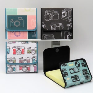 May include: A collection of square notepad holders with camera designs. The holders feature various patterns, including teal, pink, and black backgrounds with white camera illustrations. One holder is open, revealing yellow sticky notes.
