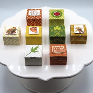 May include: Eight small square cardboard boxes in various colors including orange, yellow, green, and white, each featuring a unique autumn-themed sticker on top such as a pumpkin pie, a green leaf, or the word Thankful, arranged on a white cake stand.