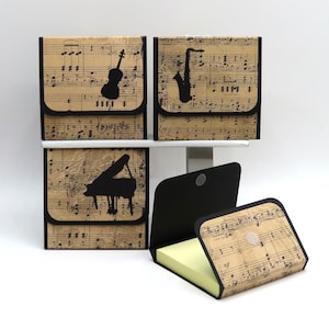 May include: Four musical-themed memo pad holders. Each holder features a black silhouette of a musical instrument: violin, saxophone, and piano. The holders are decorated with sheet music and have a black border. One holder is open, revealing yellow sticky notes.