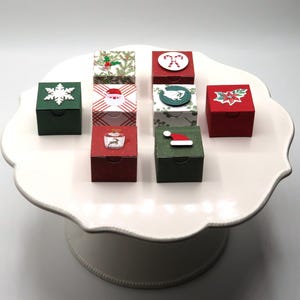 May include: Seven small square gift boxes in red and green with various holiday-themed embellishments including a white snowflake, a Santa face, a candy cane, a bird, a poinsettia, a reindeer, and a Santa hat, arranged on a white pedestal stand.