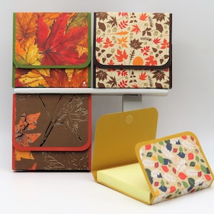 May include: Four square memo pad holders with autumn leaf designs. The top three are closed, showing colorful leaf patterns and brown trim. The bottom one is open, revealing a yellow notepad. The open holder has a leaf pattern on the cover.