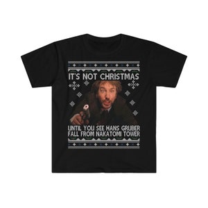 May include: Black t-shirt with a festive design. The shirt features the text "IT'S NOT CHRISTMAS" above an image of a man holding a gun, with the text "UNTIL YOU SEE HANS GRUBER FALL FROM NAKATOMI TOWER" below.