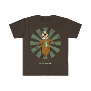 Pode incluir: Uma camiseta marrom com um gráfico de estilo retrô do Yogi Bear com um boné de piloto verde e uma echarpe. O gráfico é cercado por um design de raios de sol verde e azul. O texto "Yogi Bear" é impresso abaixo do gráfico em verde e branco, com os caracteres japoneses para "Yogi Bear" acima.