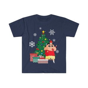 May include: Navy blue t-shirt with a cartoon character wearing a Santa hat, standing beside a decorated Christmas tree and wrapped presents. White snowflakes are scattered around the tree. The character wears a red shirt and yellow shorts.