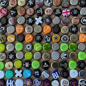 May include: A selection of beer bottle caps featuring diverse branding and designs, including logos and text such as "London Pride," "Beck's," and "San Miguel." The caps are displayed on a textured, beige surface.