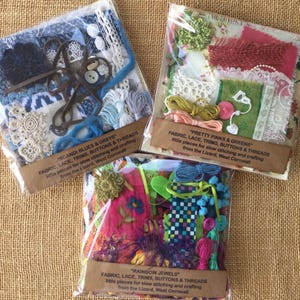 May include: Three clear plastic packets containing assorted craft supplies including fabric scraps, lace, ribbons, buttons, and thread. Each packet features a brown paper label with text identifying the colour theme as Scandi Blues & Greys, Pretty Pinks & Greens, or Rainbow Jewels, along with the origin from the Lizard, West Cornwall.