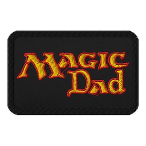 May include: Black embroidered patch with the words "MAGIC Dad" in yellow and red thread. The patch has a black border and is rectangular in shape. The text is stylized with a fantasy theme.