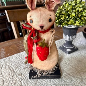 May include: A handcrafted bunny figurine with a distressed aesthetic. The bunny is beige with large ears, black eyes, and a red nose. It holds a red strawberry and is adorned with a red ribbon. The figurine sits on a black base.