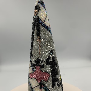May include: A silver Christmas tree ornament with a bell on top. The tree is covered in silver sequins and features a design of dancing figures in black and pink. The tree is approximately 8 inches tall.