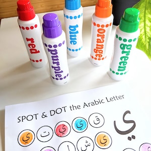 May include: A set of five colorful dot markers with the words 'red', 'purple', 'blue', 'orange', and 'green' written on them. The markers are arranged in a semi-circle on a white surface. A worksheet with the Arabic letter 'y' and the words 'SPOT & DOT the Arabic Letter' is visible in the background.