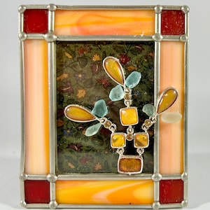 May include: A stained glass picture frame with a peach and red border. The frame contains a green fabric background with a decorative design of amber and blue glass pieces.