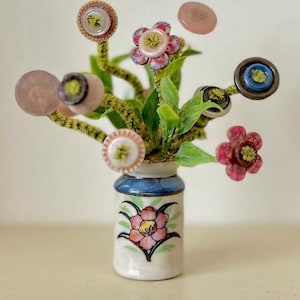 May include: A small ceramic vase with a painted pink flower design holds a bouquet of artificial green stems topped with various colorful buttons. The buttons are round, featuring shades of pink, blue, and brown, with yellow centers.