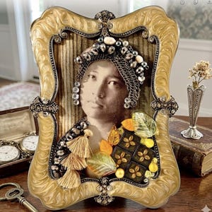 May include: Ornate gold-toned picture frame with a vintage photograph. The frame features intricate detailing, including rhinestones and embellishments. The photograph depicts a woman wearing a jeweled headpiece and decorative elements. A small vase with dried flowers is visible.
