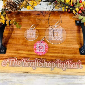 May include: Three clear acrylic keychains with a pink glitter keychain in the center. The keychains feature a QR code design. The keychains are displayed on a wooden surface with the text "@The KraftShop By Kari" in pink glitter.