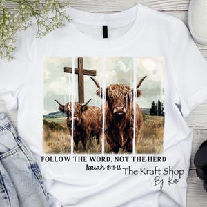 May include: White t-shirt featuring a watercolor-style image of two Highland cows with a wooden cross in the background. The text "FOLLOW THE WORD, NOT THE HERD" and "Isaiah 8:11-13" are printed below the image. The Kraft Shop By Kari.