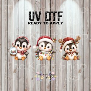 May include: Three adorable penguin designs for UV DTF transfers. Each penguin wears a festive hat and scarf, with one holding a snow globe, one with Christmas lights, and one with antlers. The text "UV DTF READY TO APPLY" is at the top.