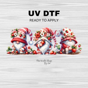 May include: Three gnomes wearing red and white hats with strawberry accents. Each gnome is holding a strawberry and has a white beard. The gnomes are standing on a wooden background with the text "UV DTF READY TO APPLY" and "The Kraft Shop By Kai" at the bottom.