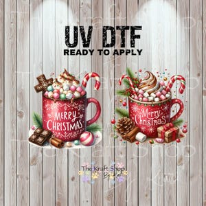 May include: Two red mugs with the text "Merry Christmas" are filled with whipped cream, marshmallows, and candy canes. The image also includes chocolate, cinnamon sticks, and the text "UV DTF READY TO APPLY".