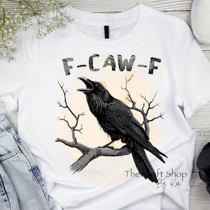 May include: White t-shirt with a black raven perched on a branch. The raven is facing left and has its beak open. The text "F-CAW-F" is above the raven. The text "The Craft Shop By Kari" is below the raven.