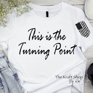 May include: White t-shirt with the text "This is the Turning Point" in black script. An American flag design is on the sleeve. The Kraft Shop by Kari is printed at the bottom.
