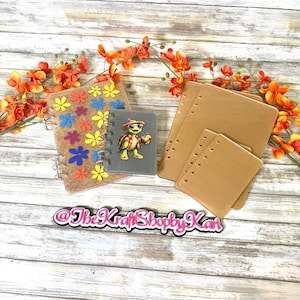 May include: A collection of stationery items, including notebooks and paper inserts, arranged on a wooden surface. One notebook features a turtle graphic, while another is decorated with colorful flower shapes. The text "@TheKraftShopyKan" is also visible.