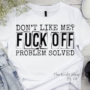 May include: White t-shirt with the text "DON'T LIKE ME? FUCK OFF PROBLEM SOLVED" in black block letters. The Kraft Shop by Kari is printed at the bottom. The shirt is displayed on a light wood surface.