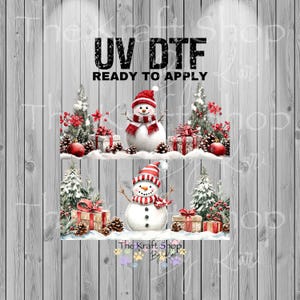 May include: A winter-themed design featuring two snowmen wearing red and white striped hats and scarves. The snowmen are surrounded by Christmas trees, gifts, pinecones, and red berries. The text "UV DTF READY TO APPLY" is at the top.