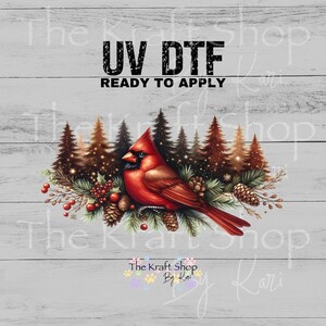May include: A vibrant red cardinal bird perched on a branch with pine cones and berries, set against a backdrop of evergreen trees. The text "UV DTF READY TO APPLY" is displayed above the bird. The Kraft Shop By Kari logo is at the bottom.