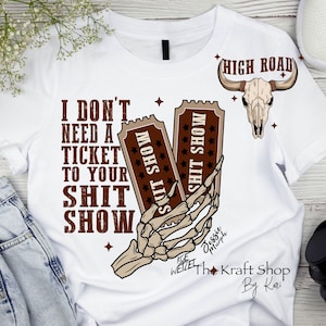 May include: White t-shirt with a humorous graphic. The design features the text "I DON'T NEED A TICKET TO YOUR SHIT SHOW" in brown, along with a skeleton hand holding two tickets. A bull skull and the words "HIGH ROAD" are also included.