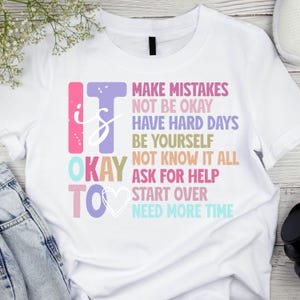 May include: White t-shirt with the words "IT IS OKAY TO" in colorful block letters. Below, the shirt lists phrases like "Make Mistakes," "Be Yourself," and "Ask for Help" in various colors.