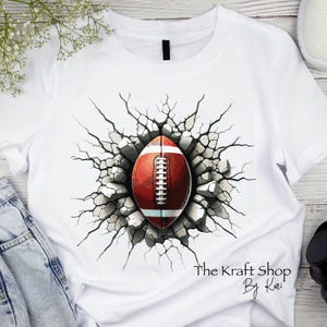 May include: White t-shirt featuring a graphic of a football bursting through a cracked wall. The football is red and white with white laces. The text "The Kraft Shop By Kari" is printed at the bottom right.