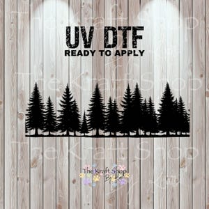 May include: Black silhouette of a forest of pine trees, with the text "UV DTF READY TO APPLY" in bold, black letters. The Kraft Shop By Kori logo is at the bottom. The image is set against a wood grain background.