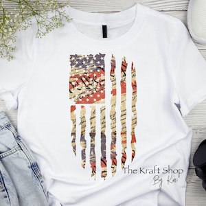 May include: White t-shirt featuring a distressed American flag design. The flag incorporates red, white, and blue colors with a vintage aesthetic. The text "The Kraft Shop By Kari" is printed at the bottom right.