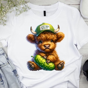 May include: White t-shirt with a cartoon illustration of a brown Highland cow wearing a green baseball cap that says "Just Dill With It." The cow is holding a green pickle.