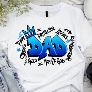 May include: White t-shirt with a blue and white graphic design that says "DAD" in large, graffiti-style letters. The graphic also includes the words "Strong", "Funny", "Hero", "Man of God", "Loving", "Hardworking", "Protector" and a crown.