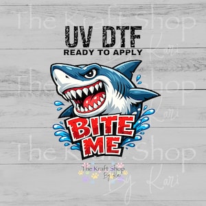 May include: A UV DTF ready to apply transfer featuring a cartoon shark with blue skin, white underbelly, and sharp teeth. The text BITE ME appears in bold red letters below the shark, surrounded by blue water splashes on a light gray background.
