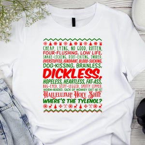 May include: White t-shirt with a red and green Christmas sweater design. The design features a list of negative adjectives and phrases, including "cheap", "lying", "no good", "rotten", "four-flushing", "low life", "dickless", "hopeless", "heartless", "fat-ass", "bug-eyed", "stiff-legged", "spotty lipped", "worm-headed", "sack of monkey shit he is!", "hallelujah!", "holy shit!", and "where's the Tylenol?"