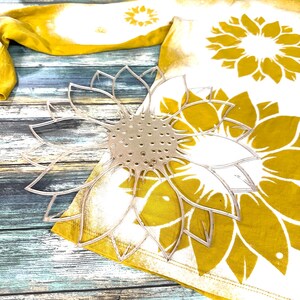 May include: A white t-shirt with a yellow and white sunflower design. The shirt is lying on a wooden surface with a clear plastic sunflower stencil on top of it.