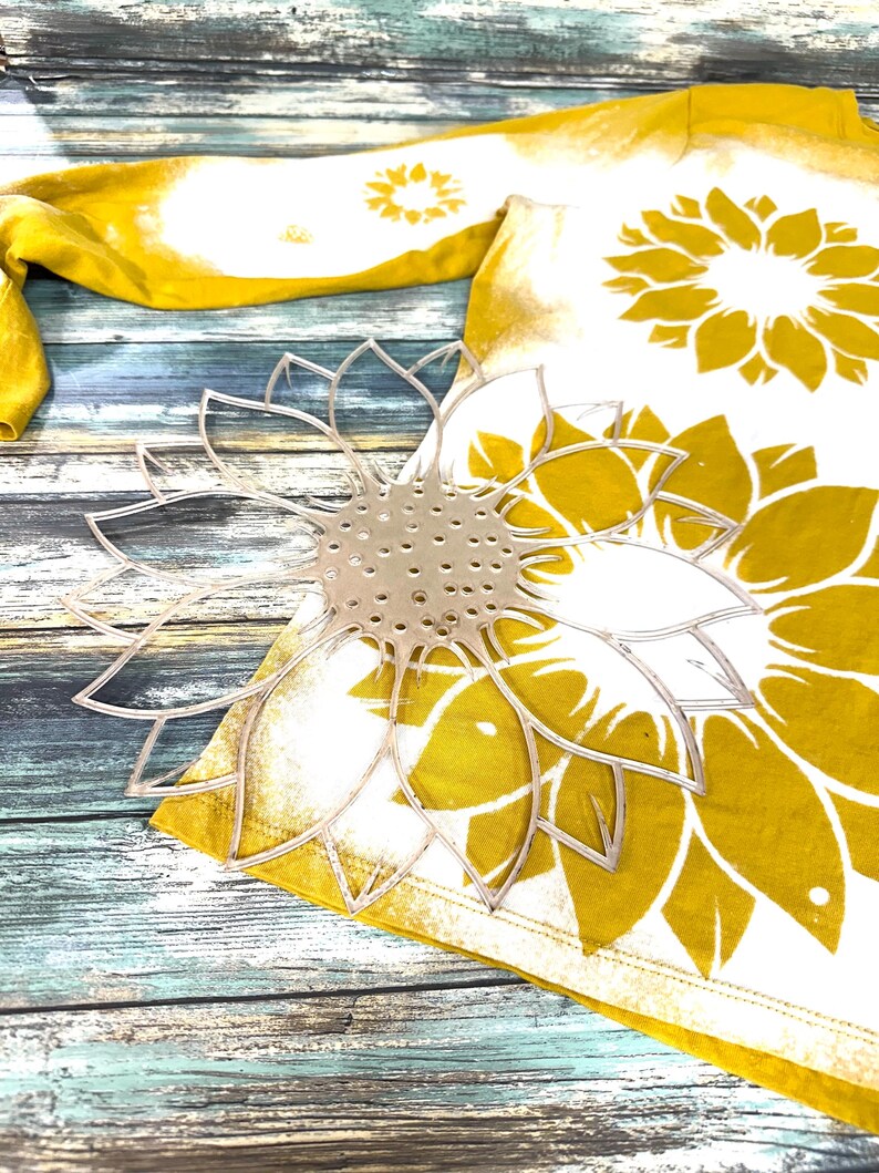 May include: A white t-shirt with a yellow and white sunflower design. The shirt is lying on a wooden surface with a clear plastic sunflower stencil on top of it.