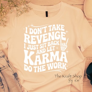 May include: A light peach-colored t-shirt with the text "I DON'T TAKE REVENGE I JUST SIT BACK AND LET KARMA DO THE WORK" in white. The design includes a peace sign made of bones. The Kraft Shop by Kari.