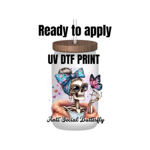 May include: A white tumbler with a wood lid and a straw. The tumbler has a UV DTF print of a skeleton wearing sunglasses and a blue and white bow. The skeleton is holding a pink butterfly. The text "Anti Social Butterfly" is printed on the tumbler.