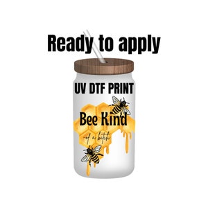 May include: A white glass tumbler with a wooden lid and straw. The tumbler features a honeycomb design with two bees and the text "Bee Kind - of a bitch". The words "Ready to apply" and "UV DTF PRINT" are also printed on the tumbler.