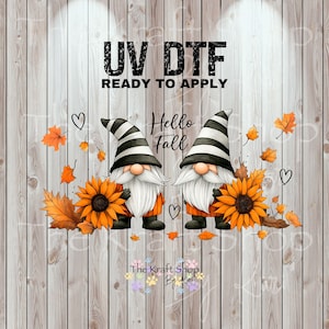 May include: UV DTF ready to apply graphic featuring two gnomes with white beards and striped hats, holding sunflowers. The design includes orange fall leaves, hearts, and the text "Hello Fall" on a wood-grain background.
