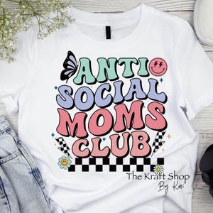 May include: White t-shirt with a colorful graphic that says "Anti Social Moms Club". The graphic includes a butterfly, a smiley face, and a checkered pattern. The text "The Kraft Shop By Kari" is at the bottom of the graphic.