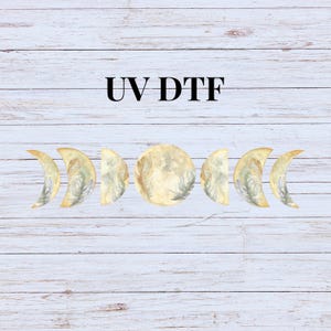May include: UV DTF design featuring a set of seven moon phases with a watercolor-like design of feathers in shades of yellow and gray on a white wood background.