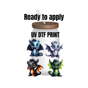 May include: A white tumbler with a wooden lid and a straw. The tumbler has four images of cartoon baby dragons in blue, gray, black, and green. The text "UV DTF PRINT" is printed on the tumbler. The text "Ready to apply" is printed above the tumbler.