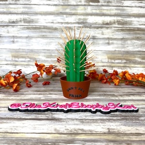 May include: A green cactus sculpture with wooden spikes in a brown pot. The pot has the text "DON'T BE A PRICK". The sculpture is on a wooden surface with orange flowers.