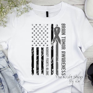 May include: White t-shirt featuring a distressed American flag design with a gray ribbon and the words "Brain Tumor Awareness" and "Nobody Fights Alone." The Kraft Shop By Kari is printed at the bottom.